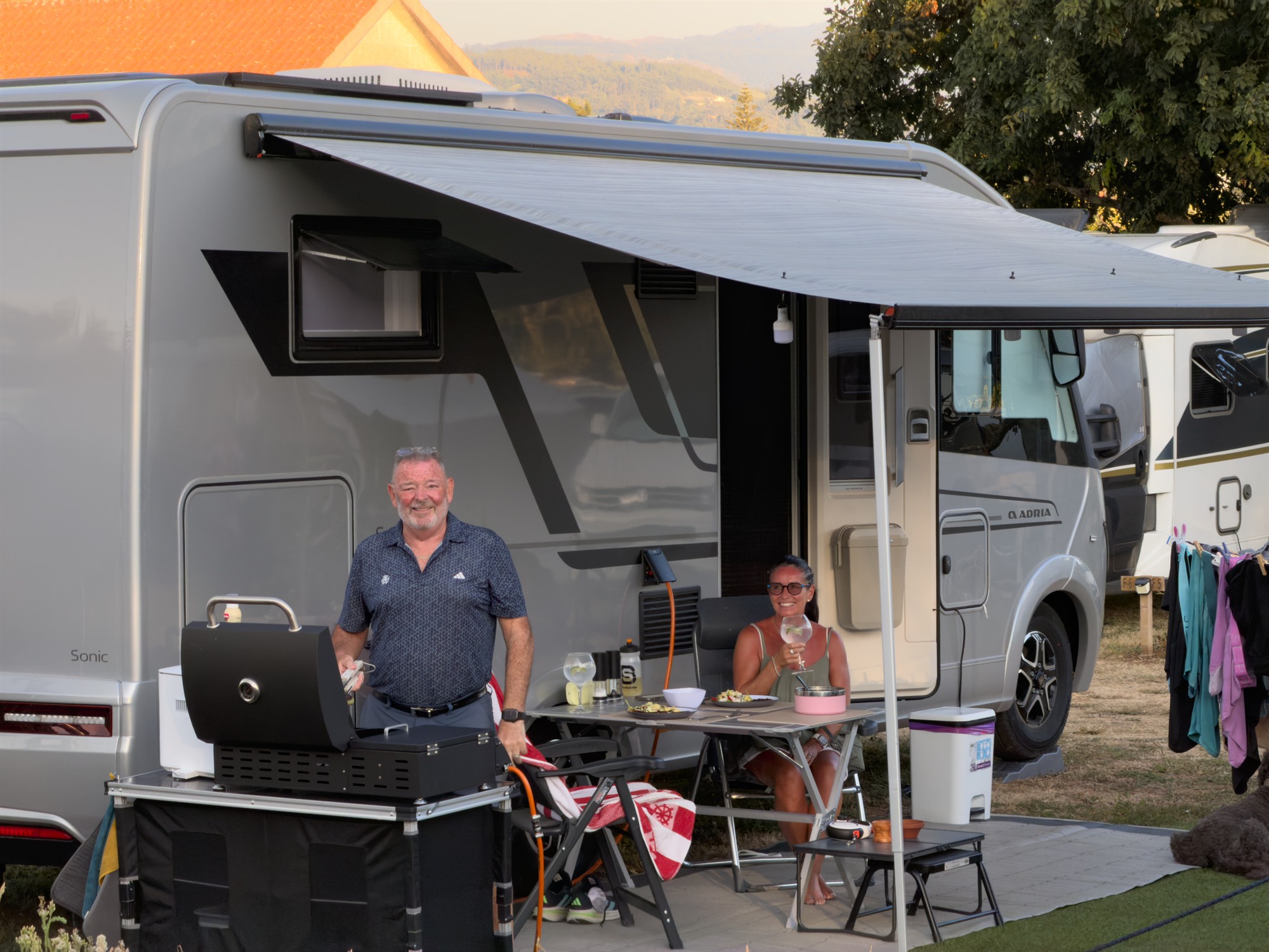Pareja de campistas comiendo bajo el toldo de su autocaravana en una plaza con alfombra de césped artificial